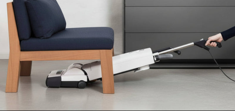 Person using a vacuum cleaner to clean under a small chair in a room with a dresser and decor.